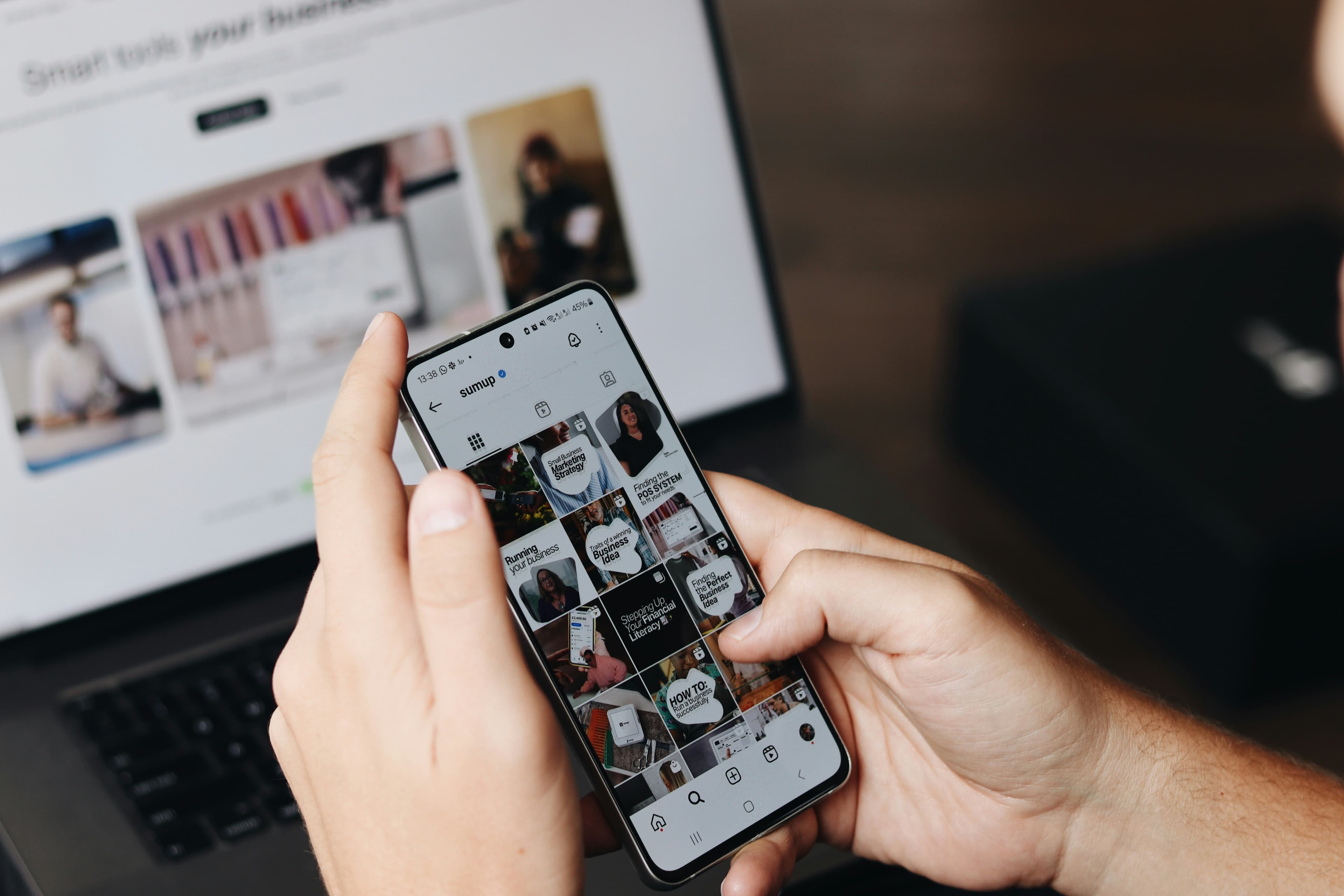 A hand scrolling an Instagram feed on a phone, a laptop open in the background