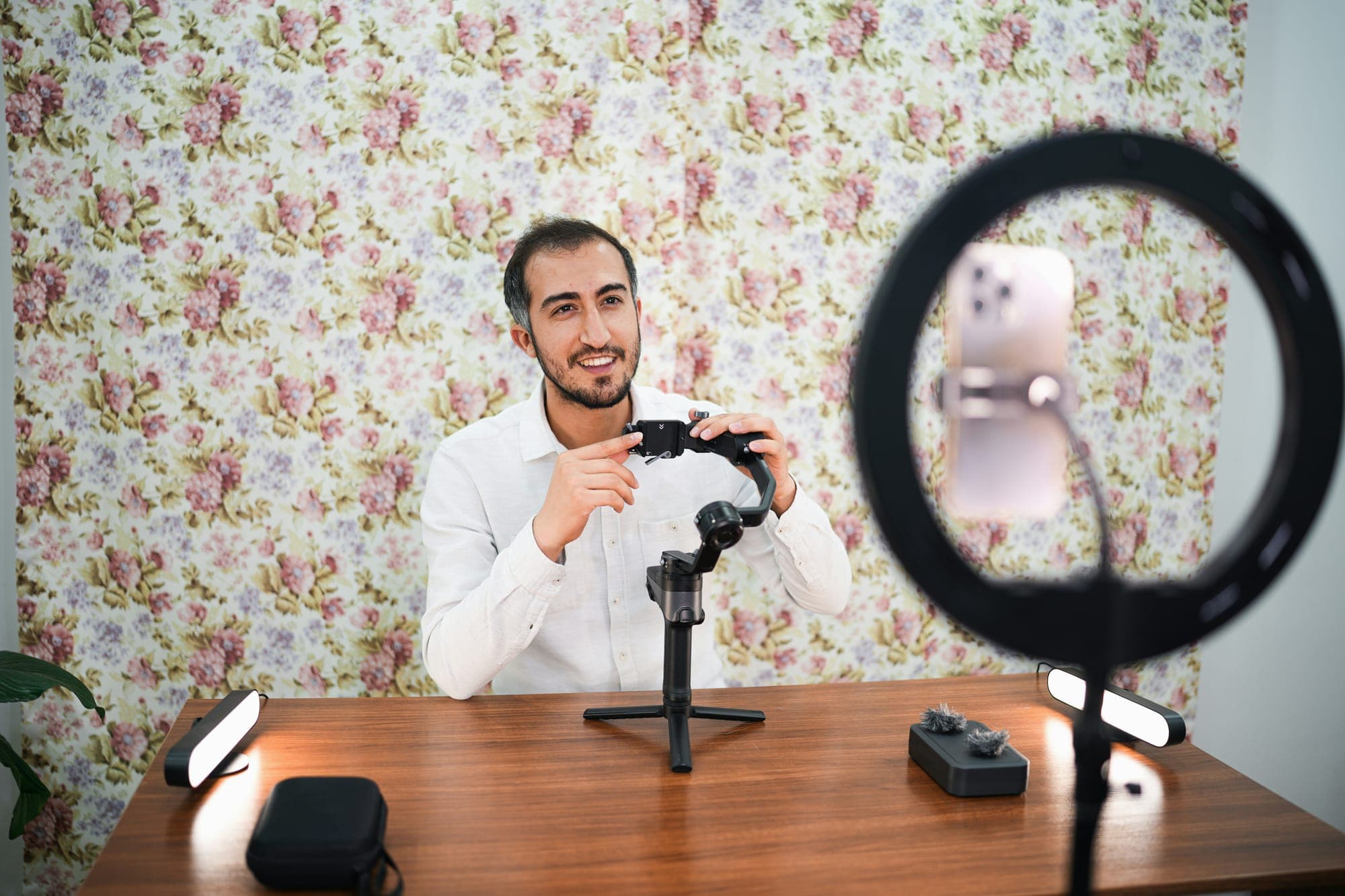 A smiling man in a white shirt holding a camera stabilizer at a wooden desk, with a ring light and a phone on a stand in the foreground