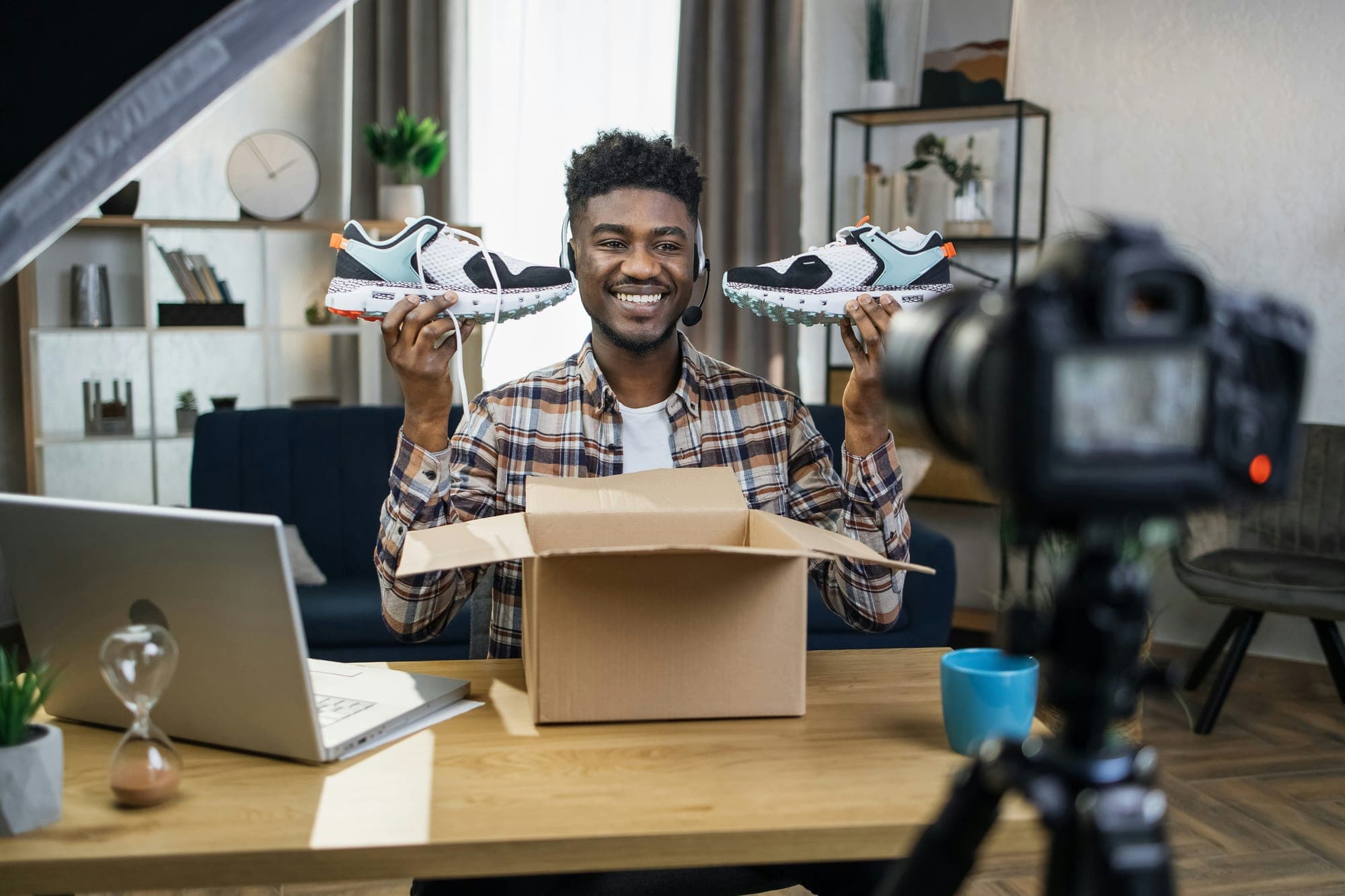 Twórca trzyma parę sneakersów nad otwartym kartonem, na biurku laptop i kamera